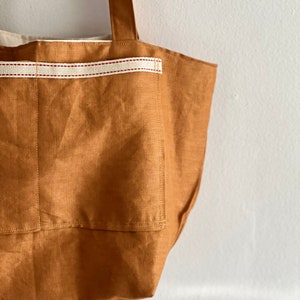 May include: A tan tote bag featuring a large exterior pocket accented with a horizontal cream-colored webbing strap that has two thin red stripes running through the center. The bag includes a sturdy shoulder strap and a light-colored interior lining.