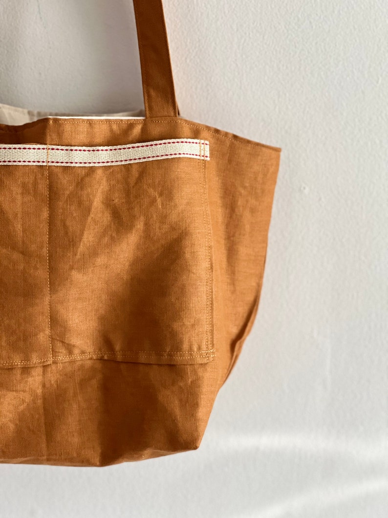 May include: A tan tote bag featuring a large exterior pocket accented with a horizontal cream-colored webbing strap that has two thin red stripes running through the center. The bag includes a sturdy shoulder strap and a light-colored interior lining.