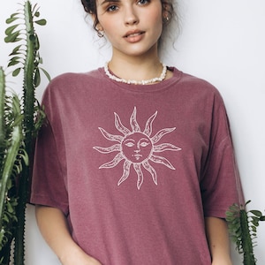 May include: A mauve oversized t-shirt with a white sun graphic and the text "Berry" printed on the bottom. The model is wearing a pearl necklace.