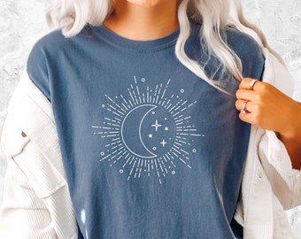 Op de afbeelding: Een blauw T-shirt met een witte afbeelding van een halve maan met sterren en zonnestralen. Het shirt wordt gedragen door iemand die blauwe denim shorts en een wit jack draagt.