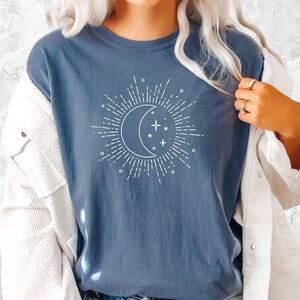 Puede incluir: Una camiseta azul con un gr&aacute;fico blanco de una luna creciente con estrellas y rayos de sol. La camiseta la lleva puesta una persona que lleva pantalones cortos de mezclilla azul y una chaqueta blanca.