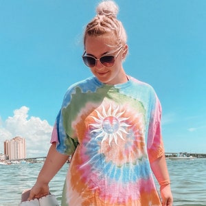 May include: A tie-dye t-shirt with a sun graphic. The shirt features a vibrant swirl of colors including blue, green, orange, and pink. The person is holding a pair of white sandals with pink soles. The background is a beach scene with blue sky and water.
