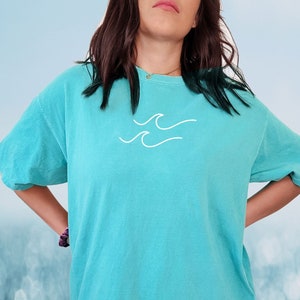 May include: A turquoise blue t-shirt with a white wave graphic. The t-shirt is oversized and has a relaxed fit. The colour of the shirt is described as "Lagoon Blue".
