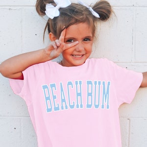 Pu&ograve; includere: Una bambina che indossa una maglietta rosa oversize con il testo "BEACH BUM" in lettere blu. La maglietta ha anche il testo "Blossom" in lettere bianche in basso.