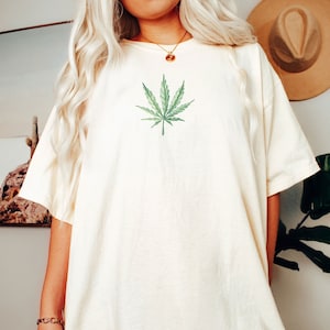 May include: An oversized white t-shirt with a green marijuana leaf graphic printed on the front.