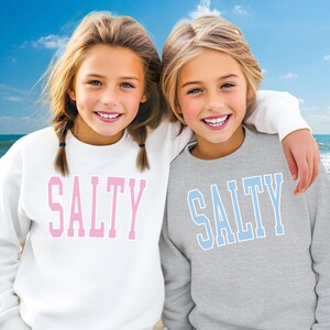 Pu&ograve; includere: Due ragazze indossano felpe abbinate grigie e bianche con la scritta "SALTY" stampata sul davanti. La ragazza a sinistra indossa una felpa bianca con lettere rosa. La ragazza a destra indossa una felpa grigia con lettere blu. Sono in piedi su una spiaggia con l'oceano sullo sfondo.