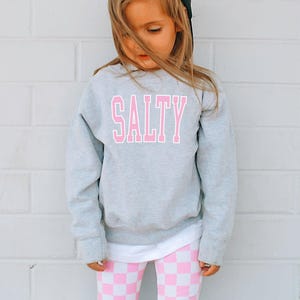 Pu&ograve; includere: Un bambino piccolo che indossa una felpa grigia con la parola "SALTY" stampata in lettere rosa. Il bambino indossa anche leggings a scacchi rosa e bianchi e un berretto da baseball nero.