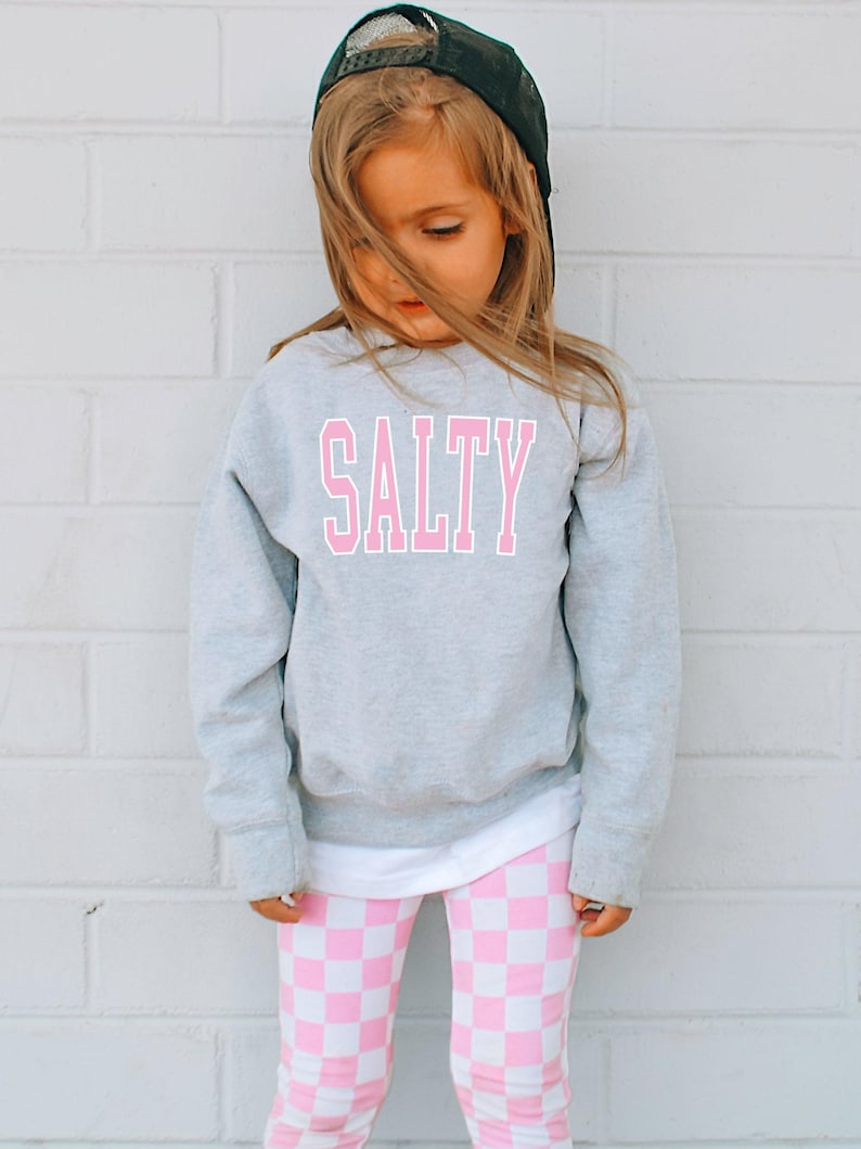 Pu&ograve; includere: Un bambino piccolo che indossa una felpa grigia con la parola "SALTY" stampata in lettere rosa. Il bambino indossa anche leggings a scacchi rosa e bianchi e un berretto da baseball nero.