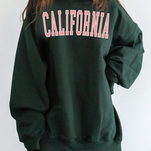 K&ouml;nnte beinhalten: Ein dunkelgr&uuml;ner Pullover mit dem Wort "CALIFORNIA" in Pink auf der Vorderseite.