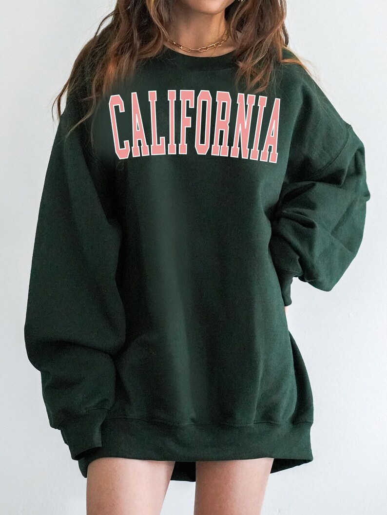K&ouml;nnte beinhalten: Ein dunkelgr&uuml;ner Pullover mit dem Wort "CALIFORNIA" in Pink auf der Vorderseite.