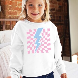 Puede incluir: Sudadera blanca con un gr&aacute;fico de rayo azul sobre un fondo de cuadros rosas.