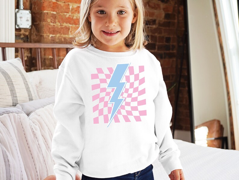 Puede incluir: Sudadera blanca con un gr&aacute;fico de rayo azul sobre un fondo de cuadros rosas.