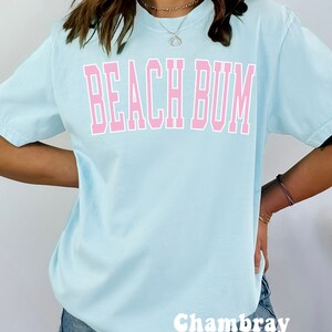 May include: A light blue t-shirt with the text "Beach Bum" in pink lettering. The shirt is made of a soft, comfortable fabric and has a relaxed fit. Chambray.