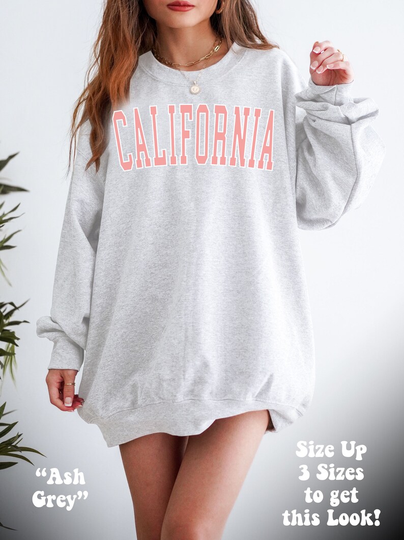 K&ouml;nnte beinhalten: Ein grauer Pullover mit dem Wort "CALIFORNIA" in Pink auf der Vorderseite. Der Pullover ist oversized und hat eine lockere Passform. Das Model tr&auml;gt den Pullover mit einer Halskette und steht vor einem wei&szlig;en Hintergrund.