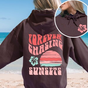 Pode incluir: Uma camisola com capuz castanho-escuro com um gr&aacute;fico nas costas com o texto "FOREVER CHASING SUNSETS" em rosa, um design de p&ocirc;r do sol &agrave;s riscas em rosa e turquesa e uma flor de hibisco turquesa. A frente inclui uma pequena flor rosa.