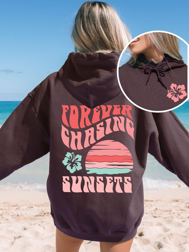 Pode incluir: Uma camisola com capuz castanho-escuro com um gr&aacute;fico nas costas com o texto "FOREVER CHASING SUNSETS" em rosa, um design de p&ocirc;r do sol &agrave;s riscas em rosa e turquesa e uma flor de hibisco turquesa. A frente inclui uma pequena flor rosa.