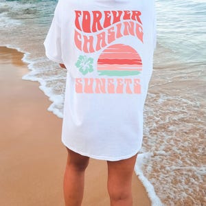 Pode incluir: Uma t-shirt branca de manga curta com um gr&aacute;fico nas costas com o texto "FOREVER CHASING SUNSETS" numa fonte ondulada de cor coral. O design inclui um p&ocirc;r do sol estilizado em tons de rosa e laranja, juntamente com uma flor de hibisco verde-clara.