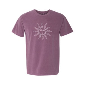 May include: A purple short-sleeved t-shirt with a white sun graphic. The sun has a face with eyes, a nose, and a mouth.