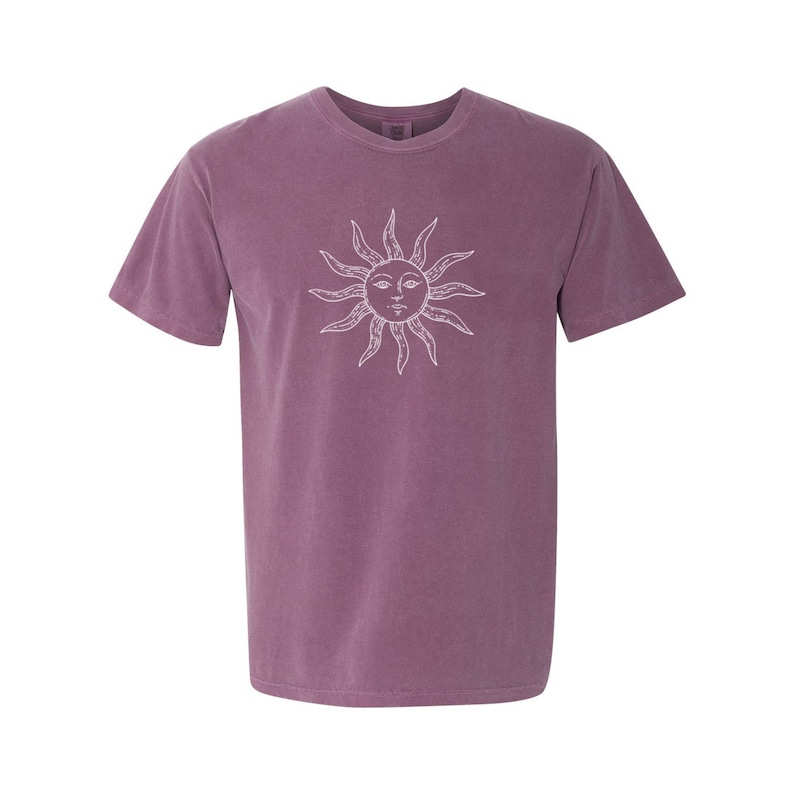 May include: A purple short-sleeved t-shirt with a white sun graphic. The sun has a face with eyes, a nose, and a mouth.