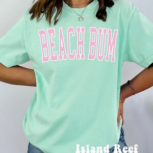 May include: A light green t-shirt with the text "Beach Bum" in pink letters. The shirt is oversized and has a relaxed fit. The brand name "Island Reef" is printed in white letters on the bottom of the shirt.