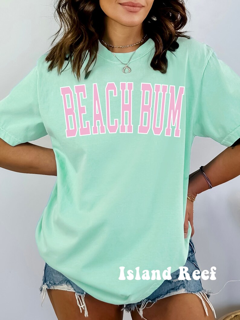May include: A light green t-shirt with the text "Beach Bum" in pink letters. The shirt is oversized and has a relaxed fit. The brand name "Island Reef" is printed in white letters on the bottom of the shirt.