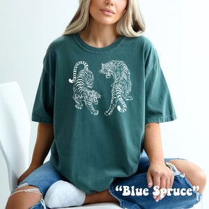 May include: A teal blue t-shirt with a white graphic of two tigers facing each other. The text "Blue Spruce" is printed on the bottom of the shirt.