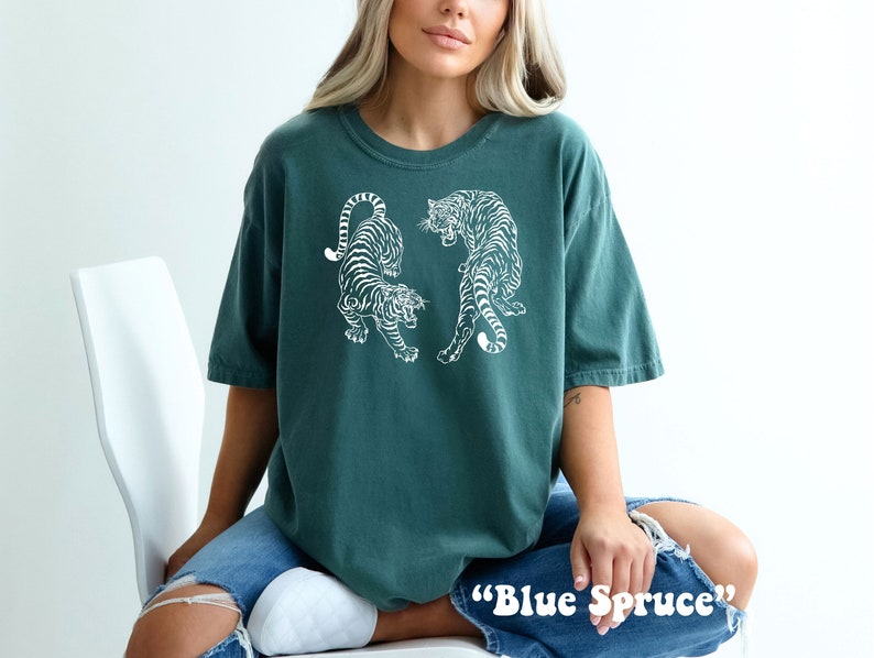 May include: A teal blue t-shirt with a white graphic of two tigers facing each other. The text "Blue Spruce" is printed on the bottom of the shirt.