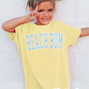 Pu&ograve; includere: Una bambina che indossa una maglietta gialla con la scritta "Beach Bum" in lettere blu. La maglietta ha le parole "Butter" in lettere bianche in basso.