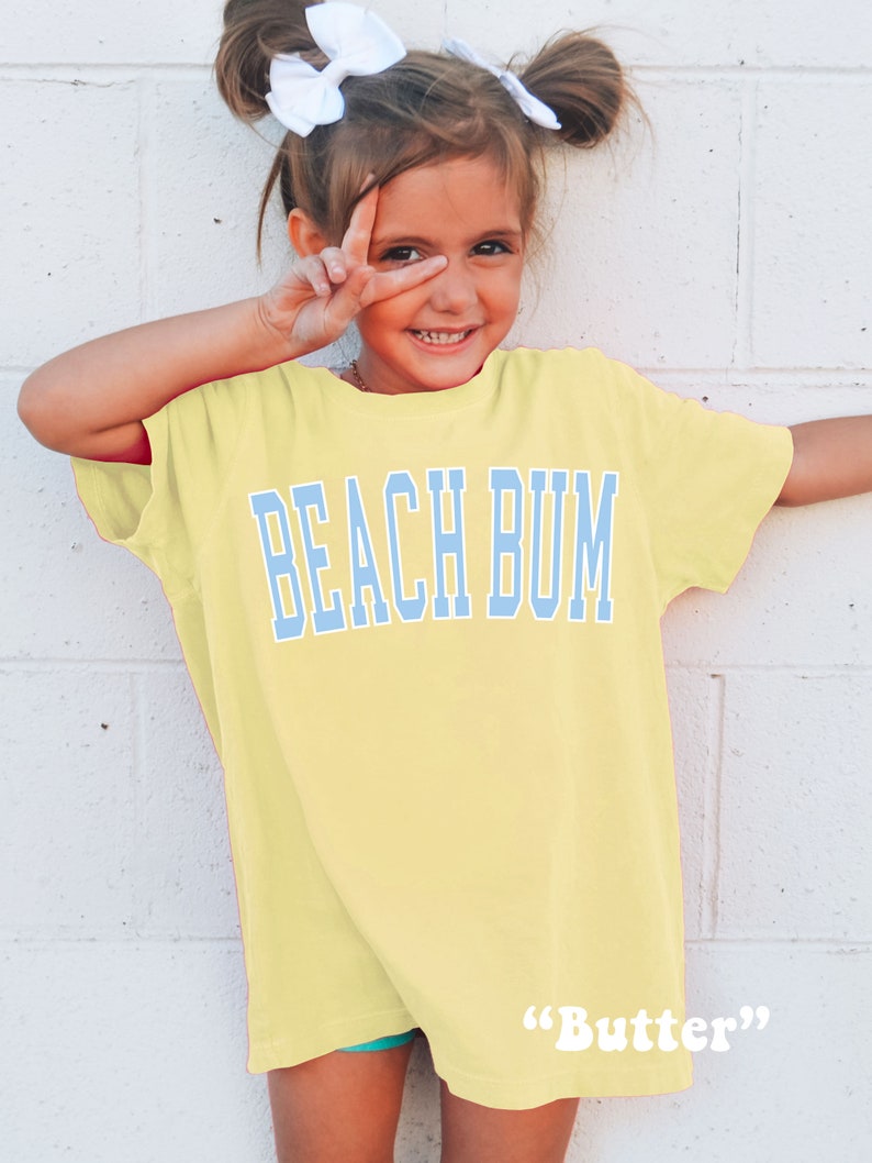 Pu&ograve; includere: Una bambina che indossa una maglietta gialla con la scritta "Beach Bum" in lettere blu. La maglietta ha le parole "Butter" in lettere bianche in basso.