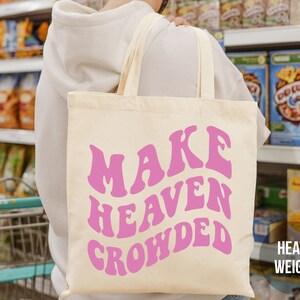 May include: A pink canvas tote bag with the text "MAKE HEAVEN CROWDED" in a retro font style. The bag is being held by a person in a grocery store.