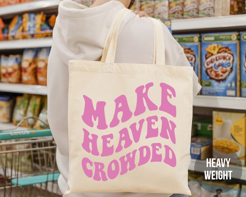 May include: A pink canvas tote bag with the text "MAKE HEAVEN CROWDED" in a retro font style. The bag is being held by a person in a grocery store.