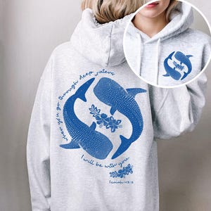 K&ouml;nnte beinhalten: Ein hellgrauer Kapuzenpullover mit blauem Aufdruck auf der R&uuml;ckseite und Vorderseite. Das R&uuml;ckendesign zeigt zwei Walhaie, florale Akzente und den Text: When you go through deep waters, I will be with you. Isaiah 43:2. Die Vorderseite hat ein kleineres, passendes Walhai-Motiv.