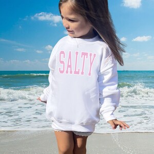 Pu&ograve; includere: Una felpa bianca con la parola "SALTY" stampata in lettere rosa. La felpa &egrave; indossata da una persona giovane in piedi su una spiaggia.