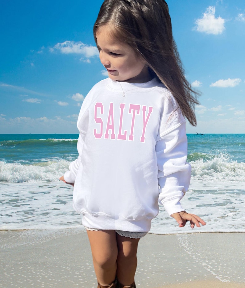 Pu&ograve; includere: Una felpa bianca con la parola "SALTY" stampata in lettere rosa. La felpa &egrave; indossata da una persona giovane in piedi su una spiaggia.