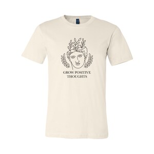 May include: A cream colored t-shirt with a black line drawing of a person with leaves growing out of their head. The text "GROW POSITIVE THOUGHTS" is printed below the drawing.
