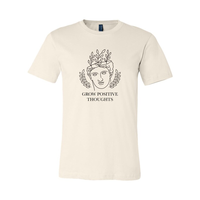May include: A cream colored t-shirt with a black line drawing of a person with leaves growing out of their head. The text "GROW POSITIVE THOUGHTS" is printed below the drawing.