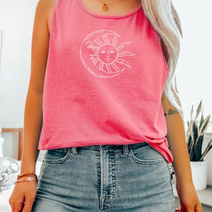 May include: A pink tank top with a white sun and moon graphic. The sleeveless top is made of a soft material and is paired with blue denim jeans. The design features a celestial theme.