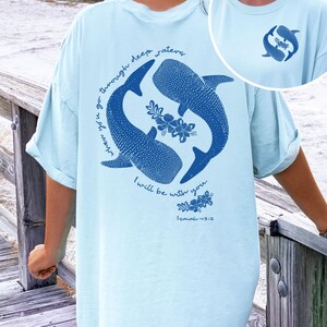 K&ouml;nnte beinhalten: Ein hellblaues T-Shirt mit einer dunkelblauen Grafik auf der R&uuml;ckseite, die zwei Walhaie zeigt, die im Kreis schwimmen, mit Blumenakzenten und dem Text "when you go through deep waters I will be with you. Isaiah 43:2". Ein kleines Logo ist vorne.