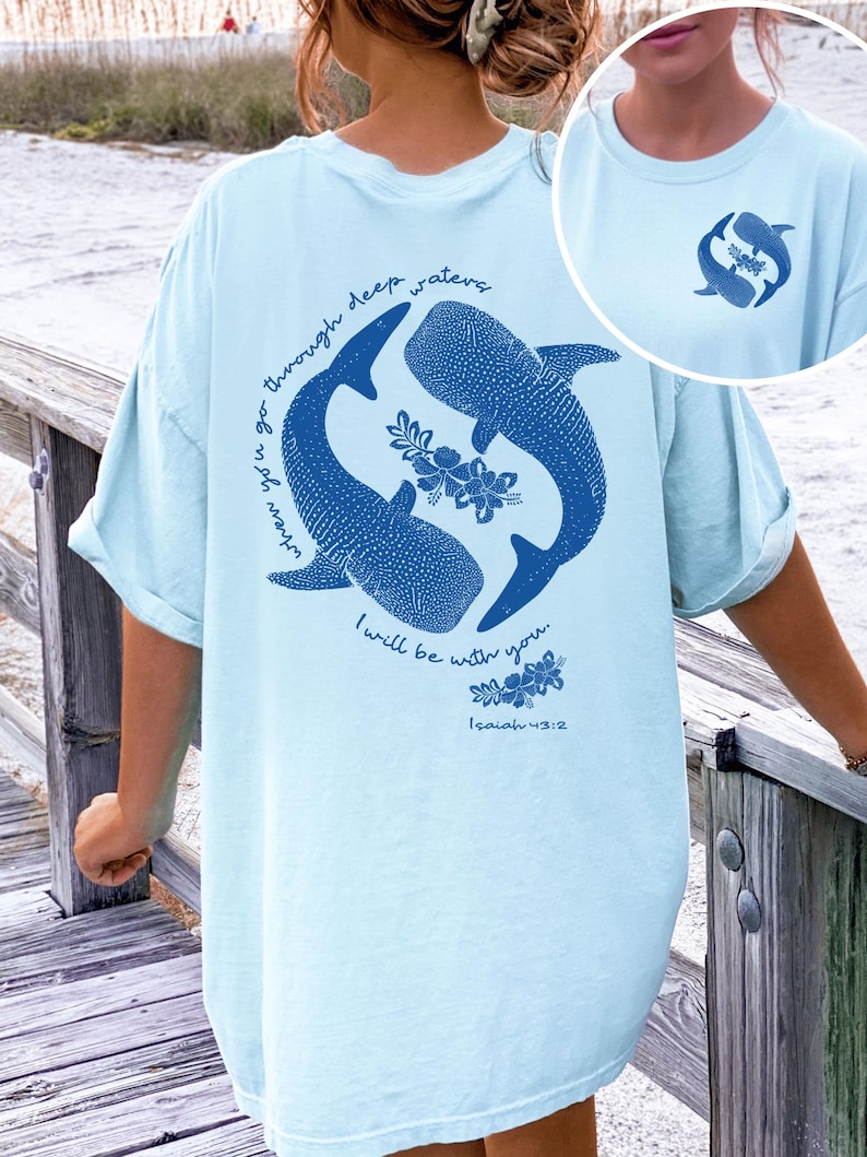 K&ouml;nnte beinhalten: Ein hellblaues T-Shirt mit einer dunkelblauen Grafik auf der R&uuml;ckseite, die zwei Walhaie zeigt, die im Kreis schwimmen, mit Blumenakzenten und dem Text "when you go through deep waters I will be with you. Isaiah 43:2". Ein kleines Logo ist vorne.