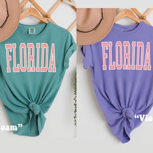Op de afbeelding: Twee T-shirts met het woord "FLORIDA" in roze op de voorkant. Het T-shirt links is turquoise en het T-shirt rechts is paars. Het turquoise T-shirt is gelabeld "Seafoam" en het paarse T-shirt is gelabeld "Violet".