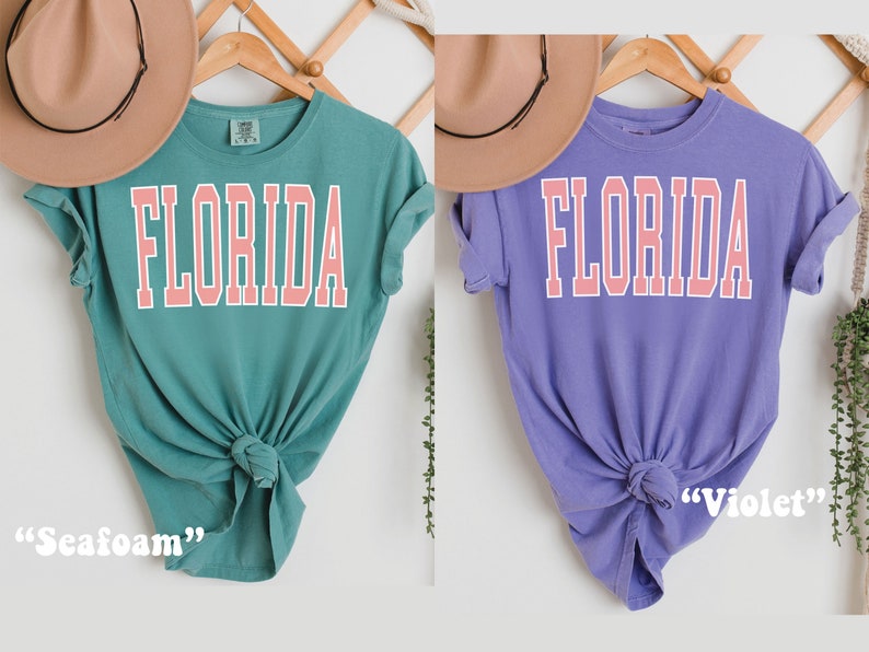 Op de afbeelding: Twee T-shirts met het woord "FLORIDA" in roze op de voorkant. Het T-shirt links is turquoise en het T-shirt rechts is paars. Het turquoise T-shirt is gelabeld "Seafoam" en het paarse T-shirt is gelabeld "Violet".