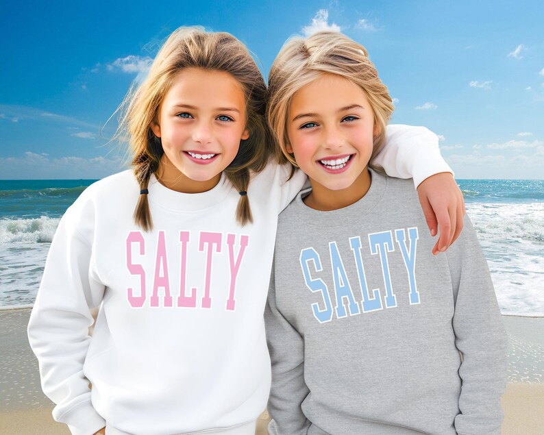 Pu&ograve; includere: Due ragazze indossano felpe abbinate grigie e bianche con la scritta "SALTY" stampata sul davanti. La ragazza a sinistra indossa una felpa bianca con lettere rosa. La ragazza a destra indossa una felpa grigia con lettere blu. Sono in piedi su una spiaggia con l'oceano sullo sfondo.
