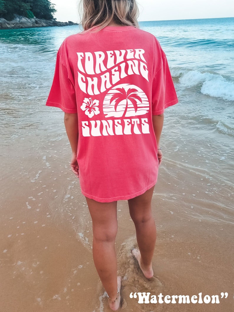 May include: A woman wearing a red t-shirt with the text "FOREVER CHASING SUNSETS" in white. The shirt features a palm tree and a hibiscus flower graphic. The text is in a retro font style. The woman is standing on a sandy beach with the ocean in the background. The caption "Watermelon" is written below the image.