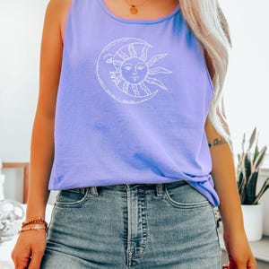 May include: A light purple tank top with a white graphic of a sun and moon design. The sun has a face and is inside a crescent moon. The tank top is paired with blue jeans.