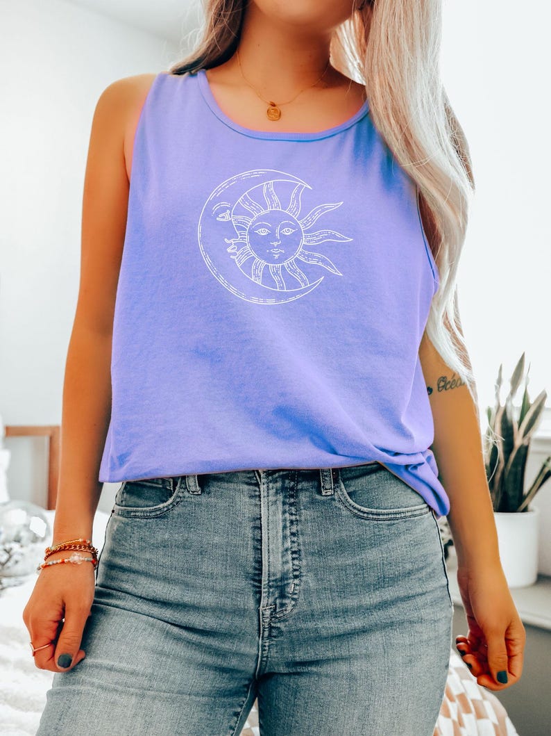 May include: A light purple tank top with a white graphic of a sun and moon design. The sun has a face and is inside a crescent moon. The tank top is paired with blue jeans.