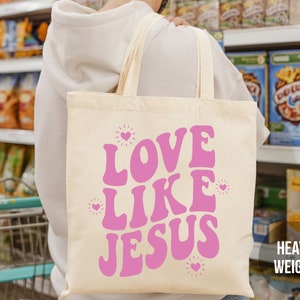 May include: A beige tote bag with a pink graphic that says "Love Like Jesus" with small hearts around the text. The bag is made of heavy weight canvas.