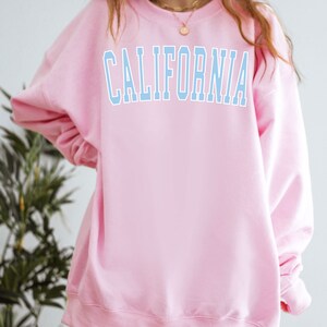 K&ouml;nnte beinhalten: Ein rosa Sweatshirt mit dem Wort "CALIFORNIA" in hellblau auf der Vorderseite.