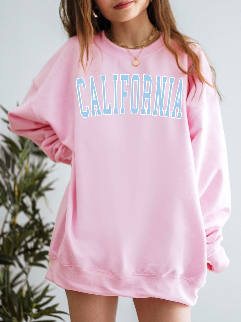 K&ouml;nnte beinhalten: Ein rosa Sweatshirt mit dem Wort "CALIFORNIA" in hellblau auf der Vorderseite.