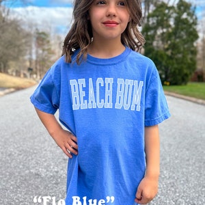 Pu&ograve; includere: Un giovane indossa una maglietta blu con la scritta "Beach Bum" in bianco. La maglietta &egrave; descritta come "Flo Blue".