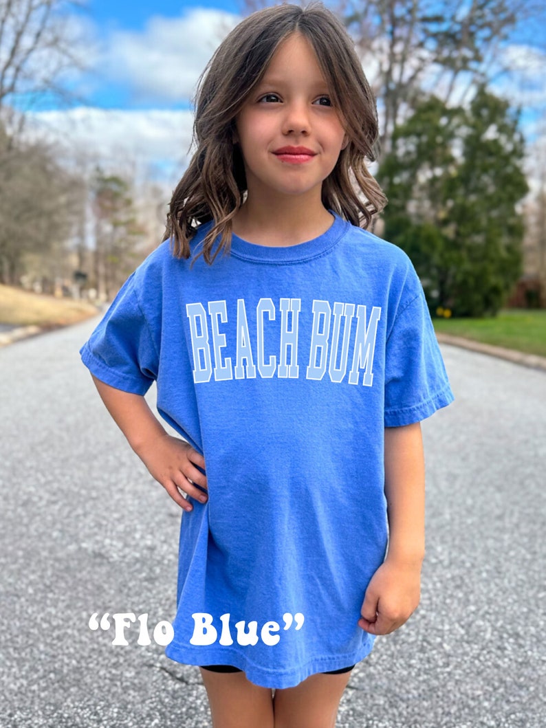 Pu&ograve; includere: Un giovane indossa una maglietta blu con la scritta "Beach Bum" in bianco. La maglietta &egrave; descritta come "Flo Blue".